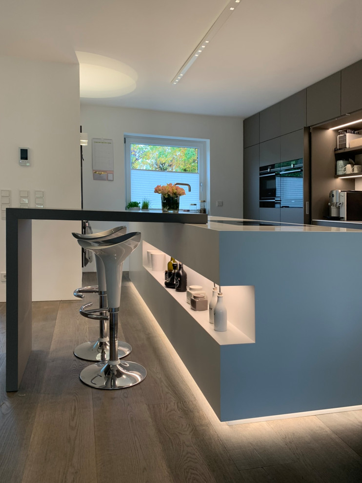 Стильный дизайн: большая кухня-гостиная в современном стиле с серыми фасадами, столешницей из акрилового камня, деревянным полом и островом - последний тренд