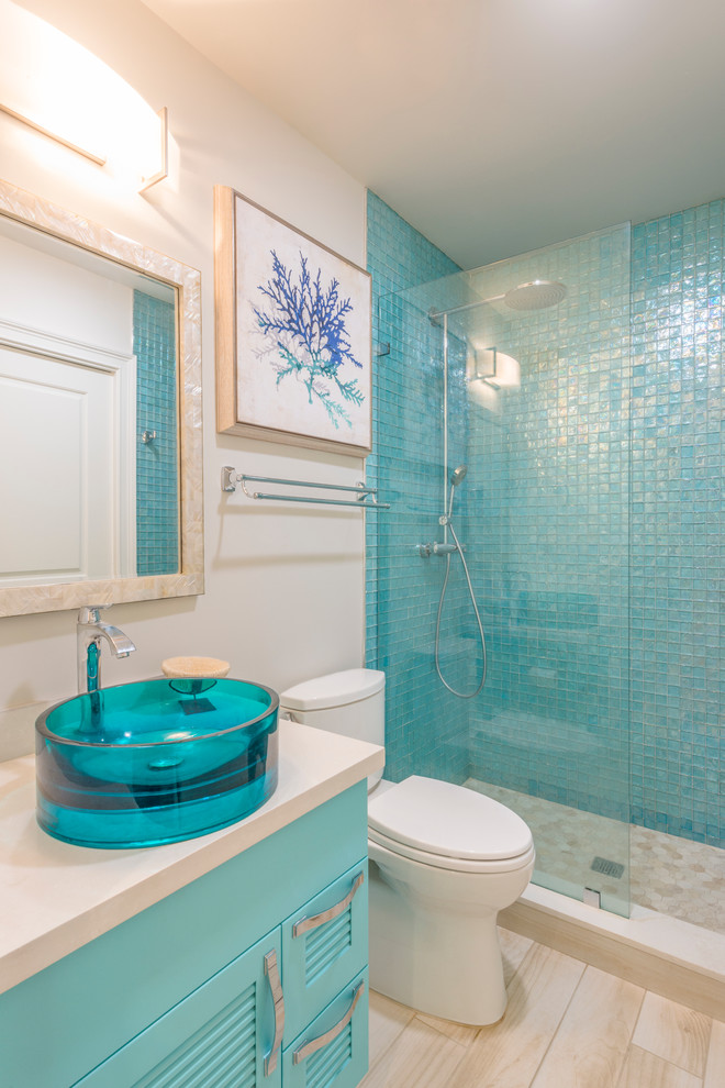 Photo of a mid-sized beach style master bathroom in Miami with louvered cabinets, blue cabinets, an alcove shower, a two-piece toilet, blue tile, glass tile, beige walls, light hardwood floors, a vessel sink, engineered quartz benchtops, beige floor, a hinged shower door and beige benchtops.