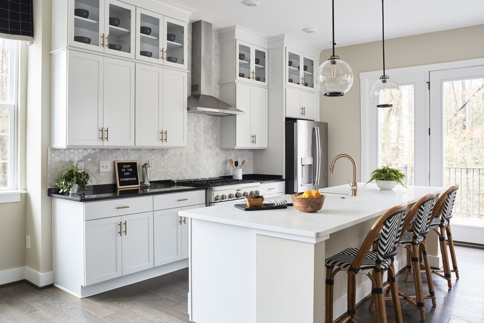 Example of a transitional galley medium tone wood floor and beige floor kitchen design with a farmhouse sink, shaker cabinets, white cabinets, gray backsplash, stainless steel appliances, an island and black countertops
