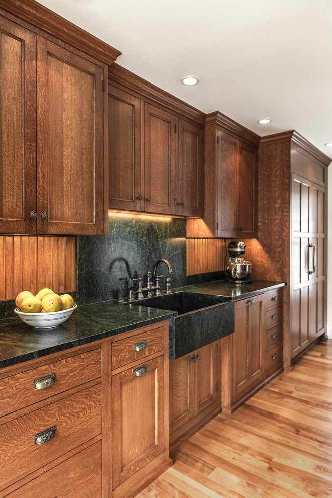 Inspiration for a mid-sized traditional l-shaped separate kitchen in Portland Maine with a farmhouse sink, recessed-panel cabinets, medium wood cabinets, soapstone benchtops, black splashback, stone slab splashback, panelled appliances, light hardwood floors and no island.