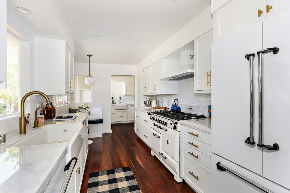 This is an example of a transitional galley separate kitchen in San Francisco with a farmhouse sink, shaker cabinets, white cabinets, white splashback, subway tile splashback, white appliances, no island, brown floor and dark hardwood floors.