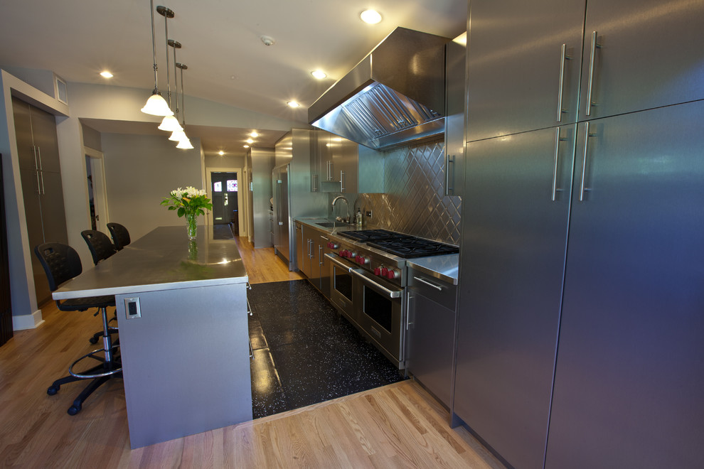 Design ideas for a large modern galley eat-in kitchen in Vancouver with a drop-in sink, flat-panel cabinets, stainless steel cabinets, stainless steel benchtops, grey splashback, glass sheet splashback, stainless steel appliances, light hardwood floors and with island.
