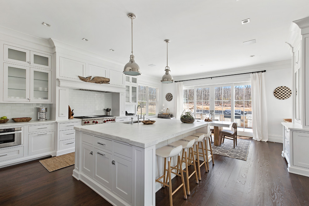 Inspiration for a mid-sized beach style l-shaped separate kitchen in New York with a farmhouse sink, shaker cabinets, white cabinets, marble benchtops, white splashback, porcelain splashback, stainless steel appliances, dark hardwood floors, with island and brown floor.