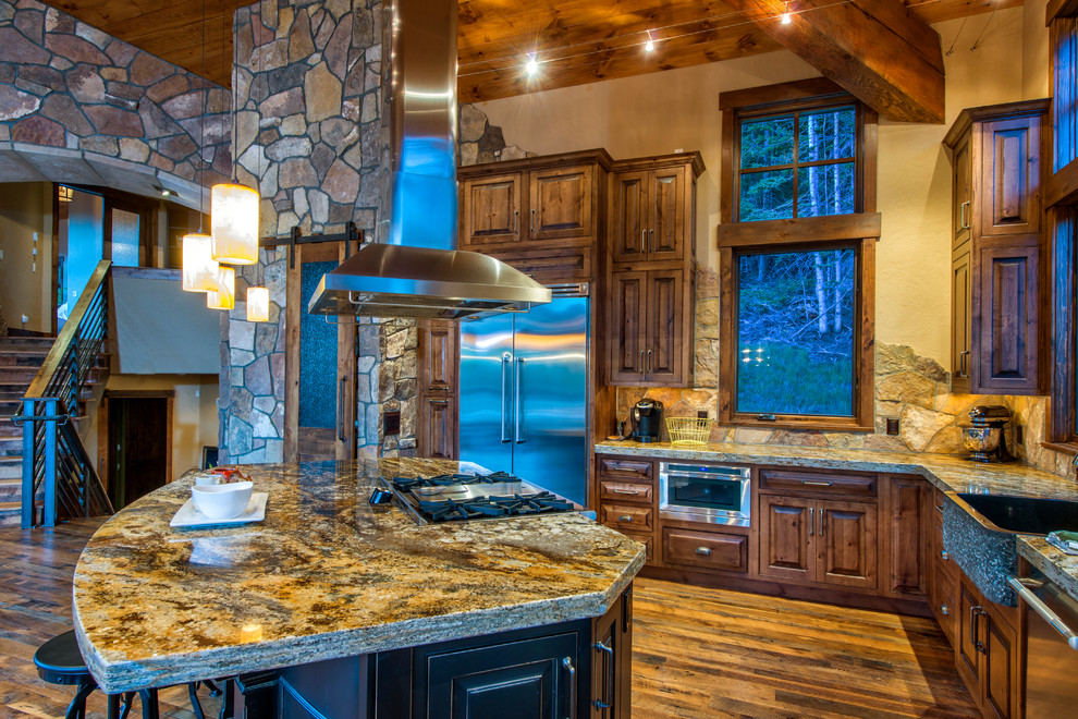 Example of a large mountain style l-shaped medium tone wood floor and brown floor open concept kitchen design in Denver with a farmhouse sink, raised-panel cabinets, medium tone wood cabinets, granite countertops, stone tile backsplash, stainless steel appliances and an island