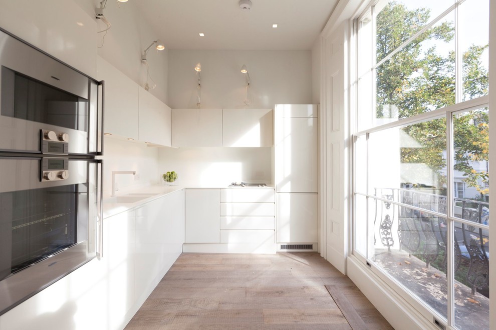 Photo of a modern l-shaped kitchen in London with a double-bowl sink, flat-panel cabinets, white cabinets, stainless steel appliances, light hardwood floors and no island.