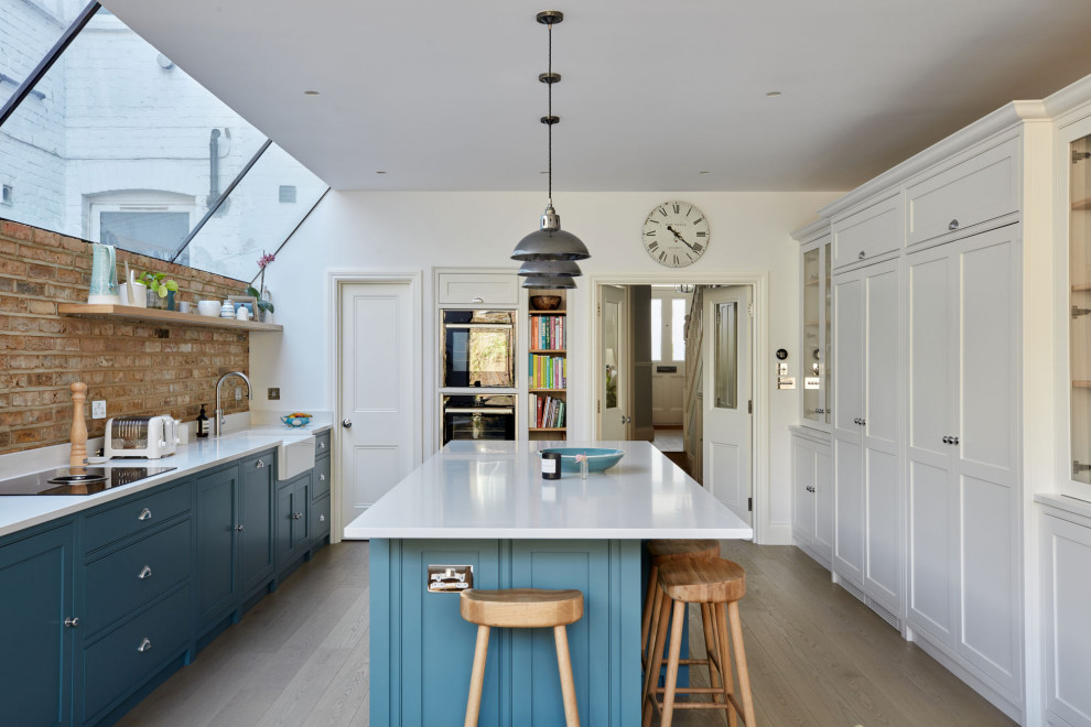 Eat-in kitchen - mid-sized contemporary single-wall medium tone wood floor and brown floor eat-in kitchen idea in London with a drop-in sink, shaker cabinets, blue cabinets, solid surface countertops, brown backsplash, brick backsplash, paneled appliances, an island and white countertops