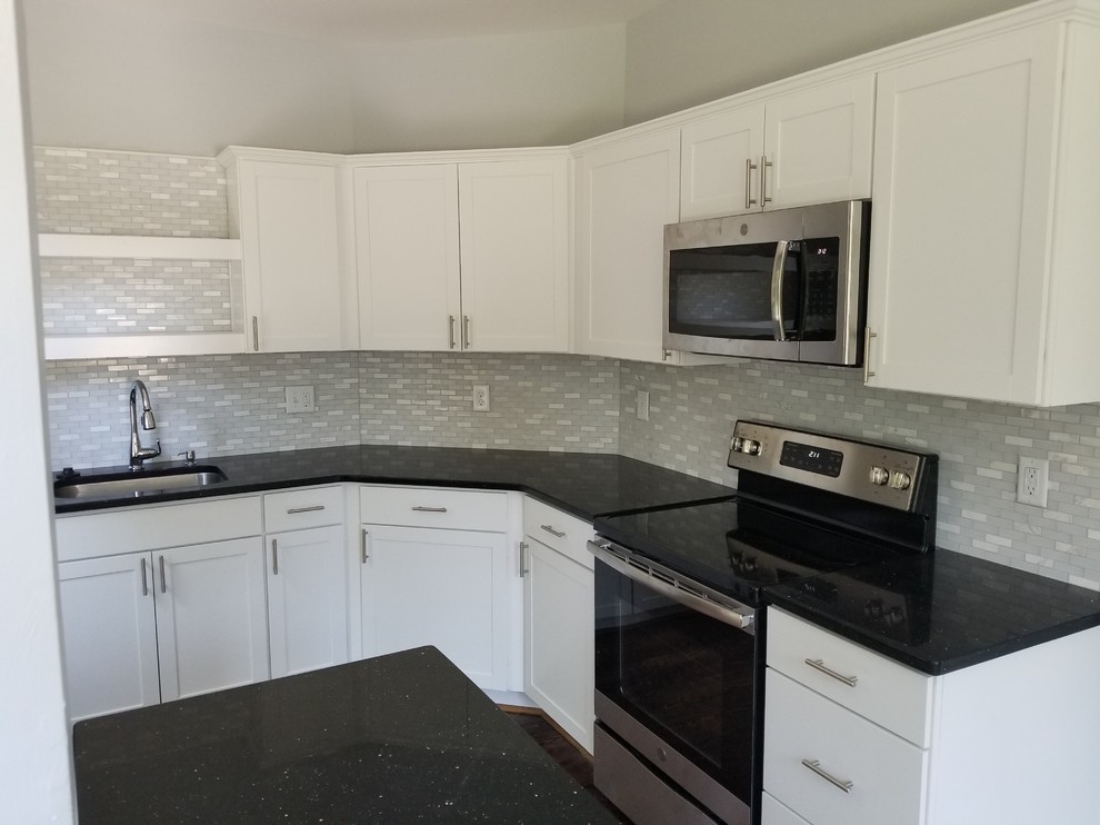 Example of a mid-sized u-shaped medium tone wood floor and multicolored floor enclosed kitchen design with an undermount sink, shaker cabinets, white cabinets, granite countertops, white backsplash, glass sheet backsplash, stainless steel appliances, an island and black countertops