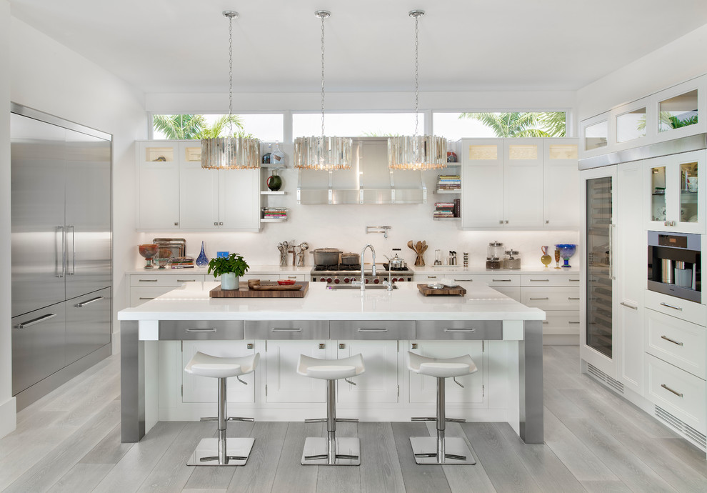 Inspiration for a beach style u-shaped kitchen in Miami with a farmhouse sink, white cabinets, white splashback, stainless steel appliances, light hardwood floors, with island, grey floor and shaker cabinets.