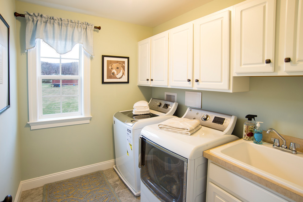 This is an example of a small traditional single-wall dedicated laundry room in Philadelphia with a drop-in sink, raised-panel cabinets, white cabinets, solid surface benchtops, green walls, slate floors, a side-by-side washer and dryer and beige benchtop.