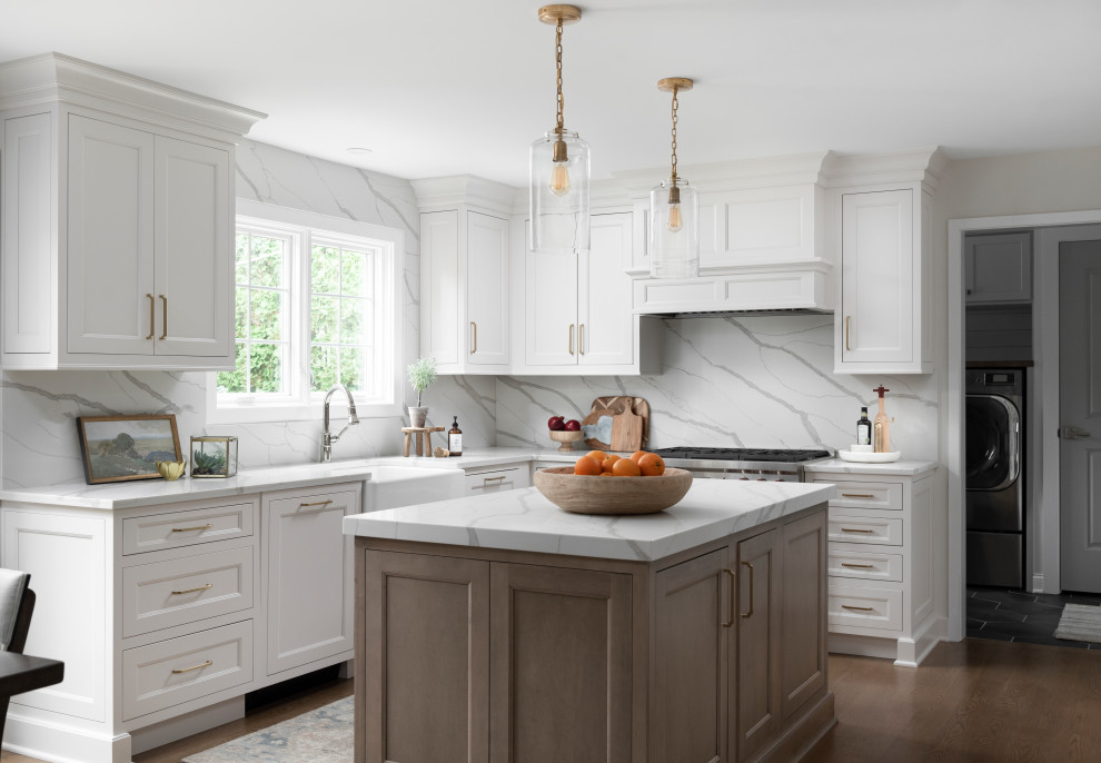 This is an example of a large traditional u-shaped kitchen in Chicago with a belfast sink, beaded cabinets, white cabinets, white splashback, stone slab splashback, stainless steel appliances, dark hardwood flooring, an island, brown floors and white worktops.