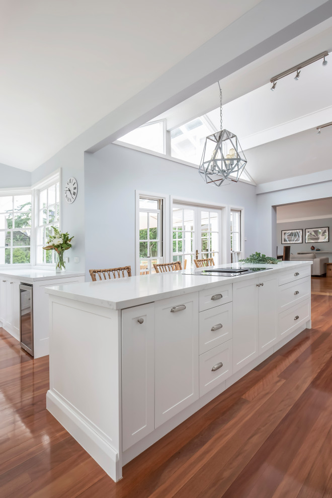 Design ideas for a large transitional u-shaped kitchen pantry in Sydney with a farmhouse sink, shaker cabinets, white cabinets, quartz benchtops, white splashback, subway tile splashback, stainless steel appliances, medium hardwood floors, multiple islands, brown floor, white benchtop and exposed beam.