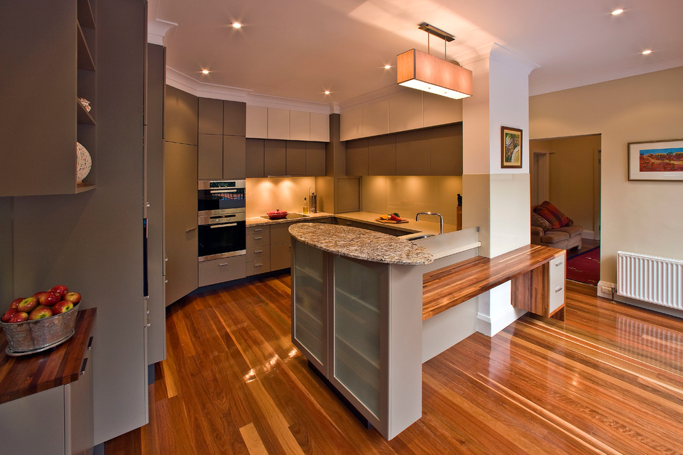This is an example of a mid-sized modern u-shaped eat-in kitchen in Melbourne with a double-bowl sink, flat-panel cabinets, grey cabinets, granite benchtops, glass sheet splashback, black appliances, medium hardwood floors and no island.