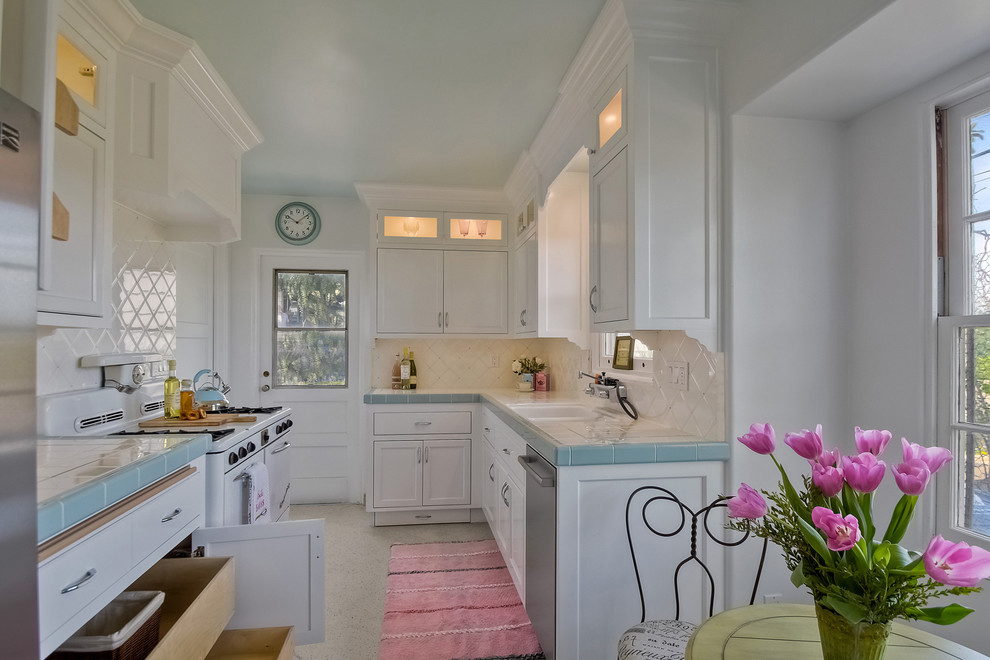 This is an example of a mid-sized transitional galley separate kitchen in San Diego with a double-bowl sink, recessed-panel cabinets, white cabinets, tile benchtops, white splashback, ceramic splashback, white appliances, terrazzo floors and no island.