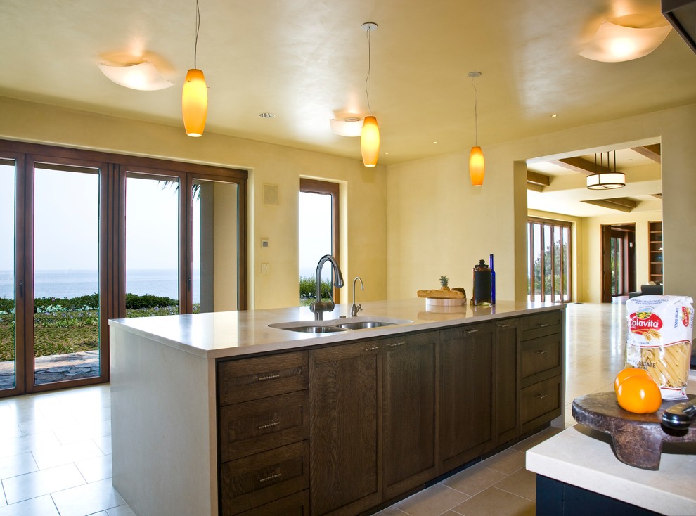 Photo of a mid-sized tropical single-wall eat-in kitchen in Santa Barbara with a double-bowl sink, shaker cabinets, black cabinets, beige splashback, stainless steel appliances, travertine floors, with island, brown floor and beige benchtop.
