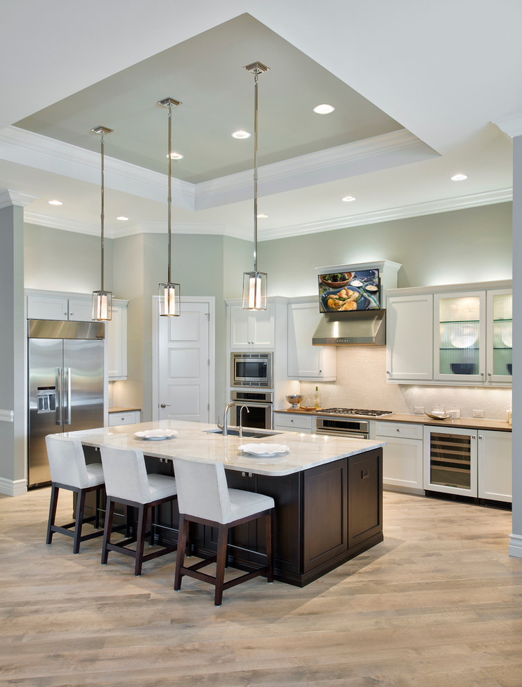 Example of a large transitional l-shaped light wood floor kitchen design with a drop-in sink, glass-front cabinets, white cabinets, granite countertops, beige backsplash, stainless steel appliances and an island