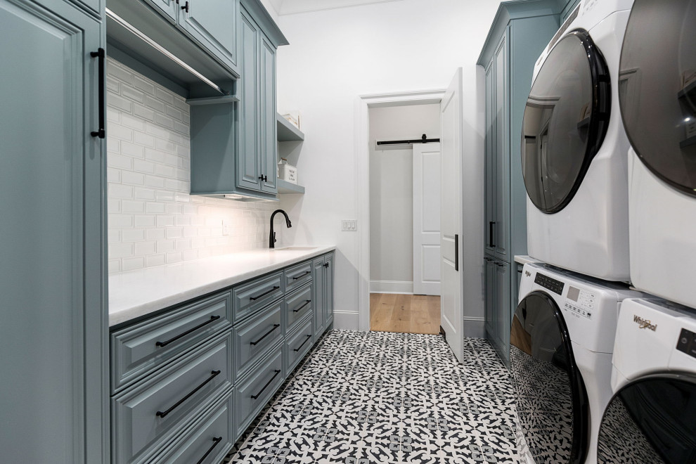 ヒューストンにある高級な広いシャビーシック調のおしゃれな洗濯室 (ll型、アンダーカウンターシンク、レイズドパネル扉のキャビネット、青いキャビネット、白いキッチンパネル、サブウェイタイルのキッチンパネル、白い壁、セラミックタイルの床、左右配置の洗濯機・乾燥機、マルチカラーの床、白いキッチンカウンター) の写真