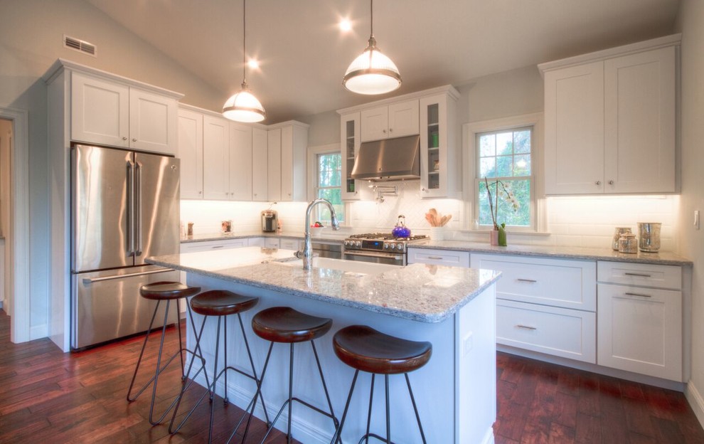 Photo of a mid-sized transitional l-shaped kitchen in Philadelphia with a farmhouse sink, shaker cabinets, white cabinets, granite benchtops, white splashback, ceramic splashback, stainless steel appliances, dark hardwood floors and with island.