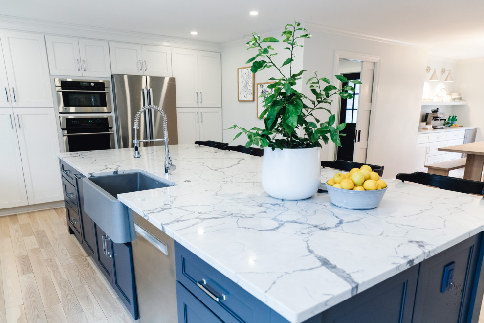 Example of a transitional light wood floor and white floor kitchen design with a farmhouse sink, flat-panel cabinets, blue cabinets, quartz countertops, blue backsplash, glass tile backsplash, stainless steel appliances, an island and white countertops