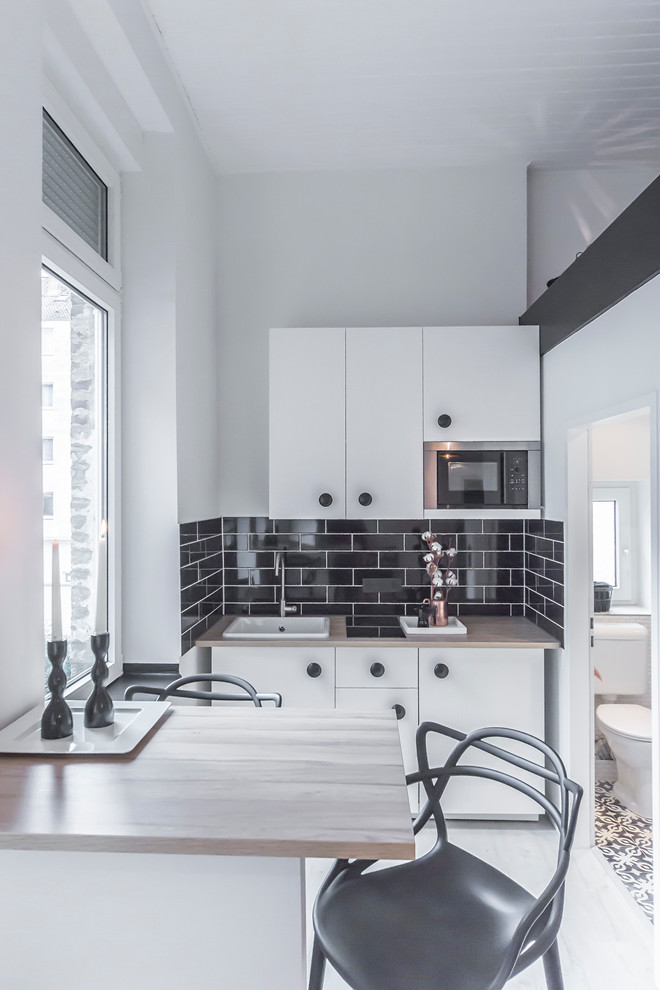 Inspiration for a small contemporary single-wall eat-in kitchen in Dusseldorf with flat-panel cabinets, white cabinets, wood benchtops, subway tile splashback, a peninsula, black splashback, stainless steel appliances, light hardwood floors and a farmhouse sink.