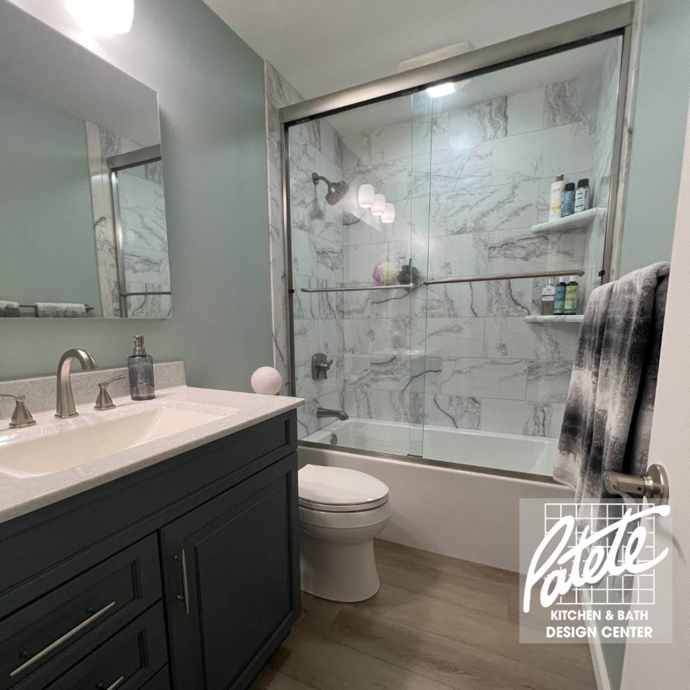 This is an example of a transitional bathroom with recessed-panel cabinets, blue cabinets, a drop-in tub, a shower/bathtub combo, a two-piece toilet, black and white tile, green walls, wood-look tile, a drop-in sink, quartzite benchtops, brown floor, a sliding shower screen, white benchtops, a single vanity and a built-in vanity.