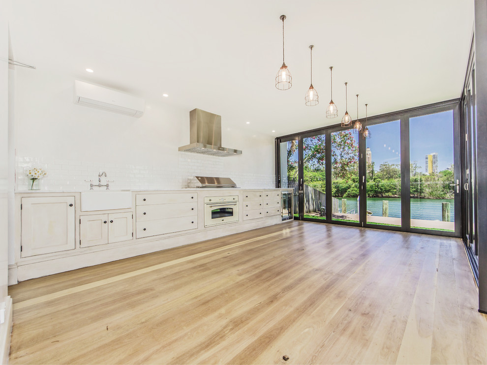 Design ideas for a large beach style single-wall eat-in kitchen in Gold Coast - Tweed with flat-panel cabinets, white cabinets, white splashback, subway tile splashback, stainless steel appliances and light hardwood floors.