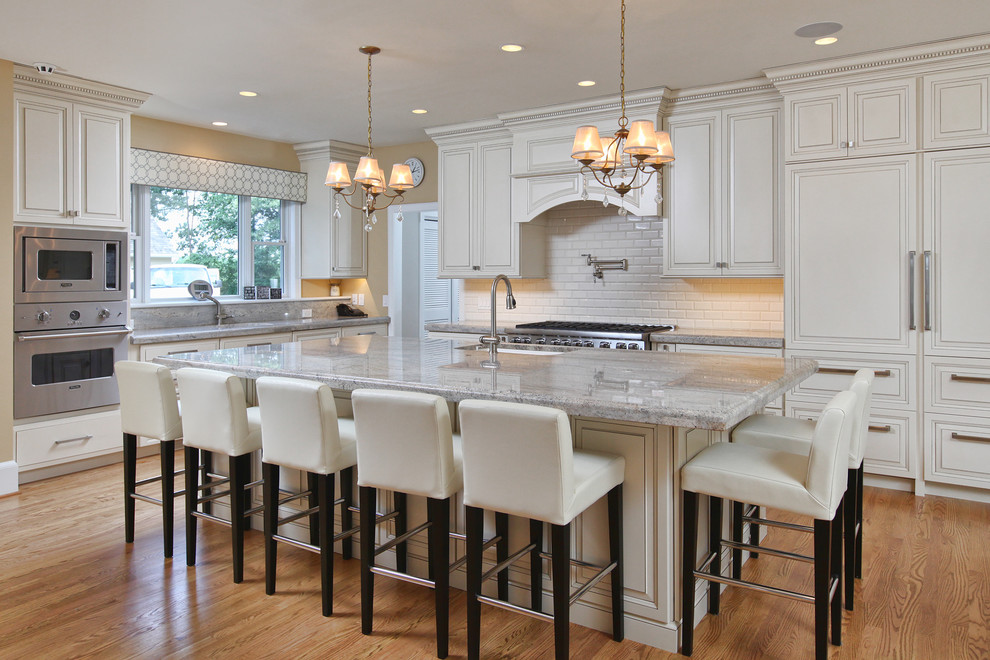 Design ideas for a large traditional l-shaped open plan kitchen in Philadelphia with raised-panel cabinets, white cabinets, white splashback, subway tile splashback, stainless steel appliances, light hardwood floors, with island, an undermount sink and granite benchtops.