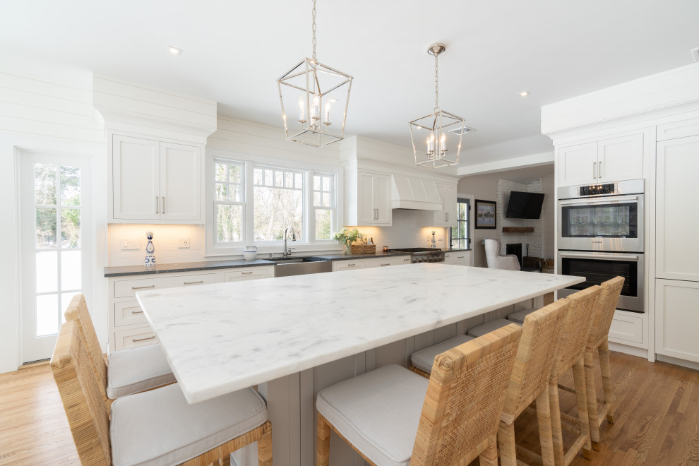 Expansive country galley eat-in kitchen in New York with a farmhouse sink, shaker cabinets, white cabinets, granite benchtops, white splashback, ceramic splashback, stainless steel appliances, medium hardwood floors, with island, brown floor and grey benchtop.