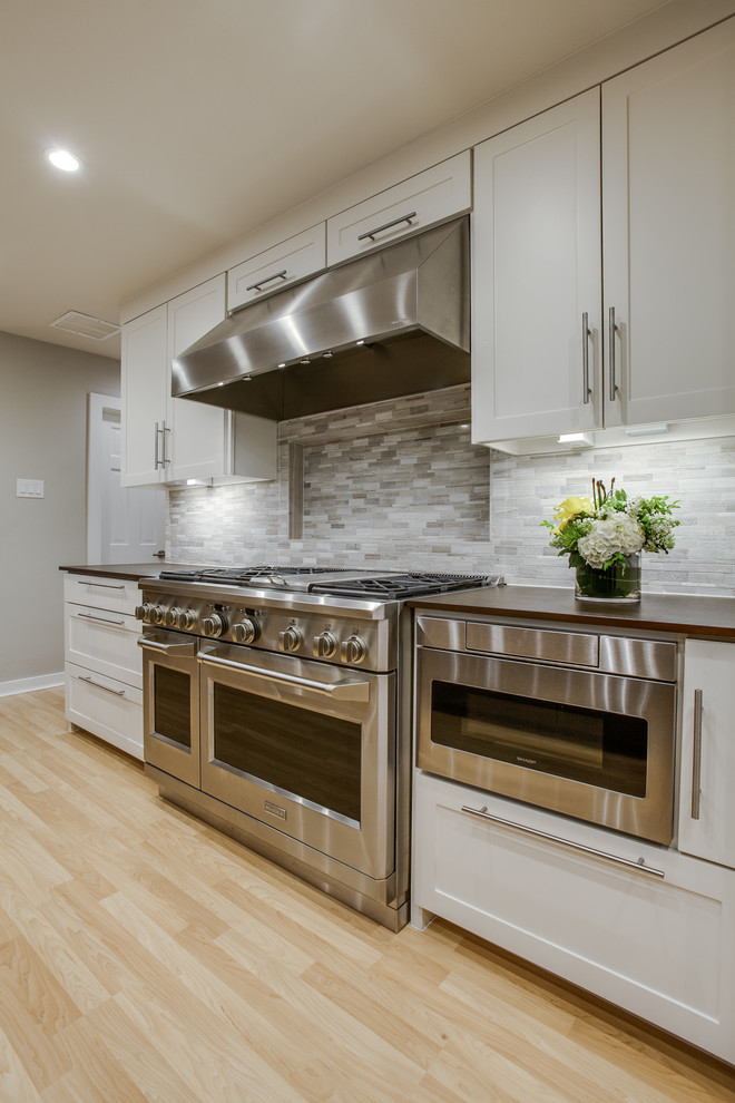 Photo of a large contemporary l-shaped open plan kitchen in Dallas with a double-bowl sink, shaker cabinets, white cabinets, quartz benchtops, grey splashback, mosaic tile splashback, stainless steel appliances, light hardwood floors and with island.