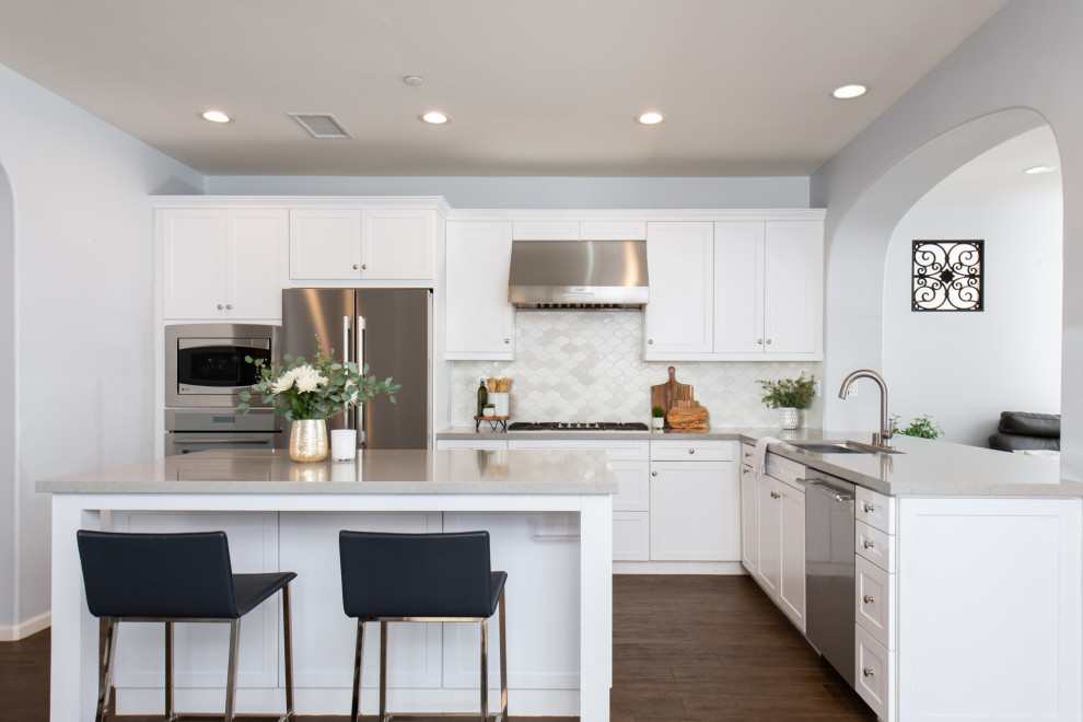 Photo of a mid-sized transitional l-shaped eat-in kitchen in Orange County with a drop-in sink, shaker cabinets, white cabinets, quartz benchtops, white splashback, porcelain splashback, stainless steel appliances, with island, brown floor and grey benchtop.