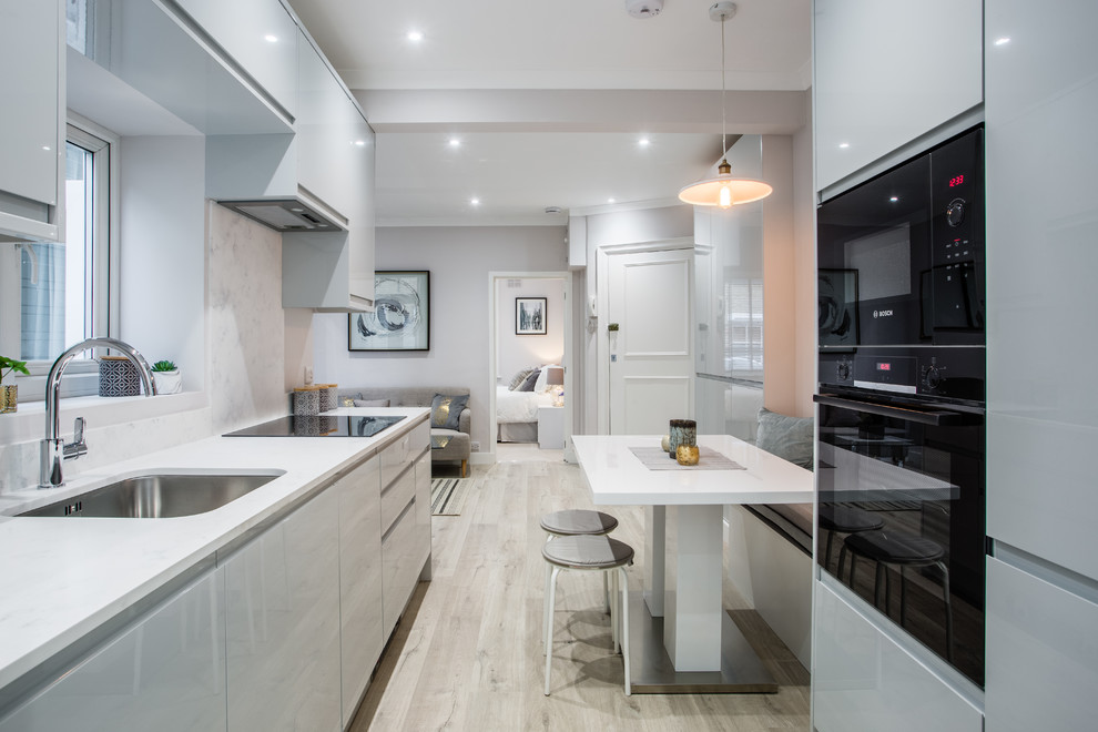 Photo of a small contemporary galley open plan kitchen in London with a drop-in sink, flat-panel cabinets, grey cabinets, solid surface benchtops, white splashback, panelled appliances, laminate floors, no island and white benchtop.