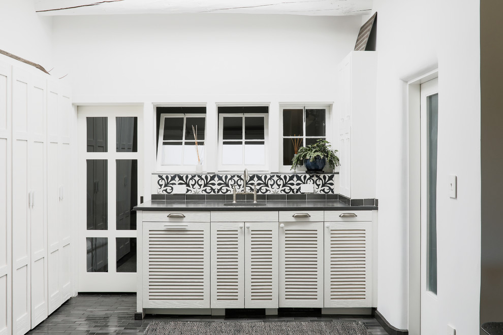 Photo of a single-wall wet bar with louvered cabinets, white cabinets, multi-coloured splashback, black floor and black benchtop.