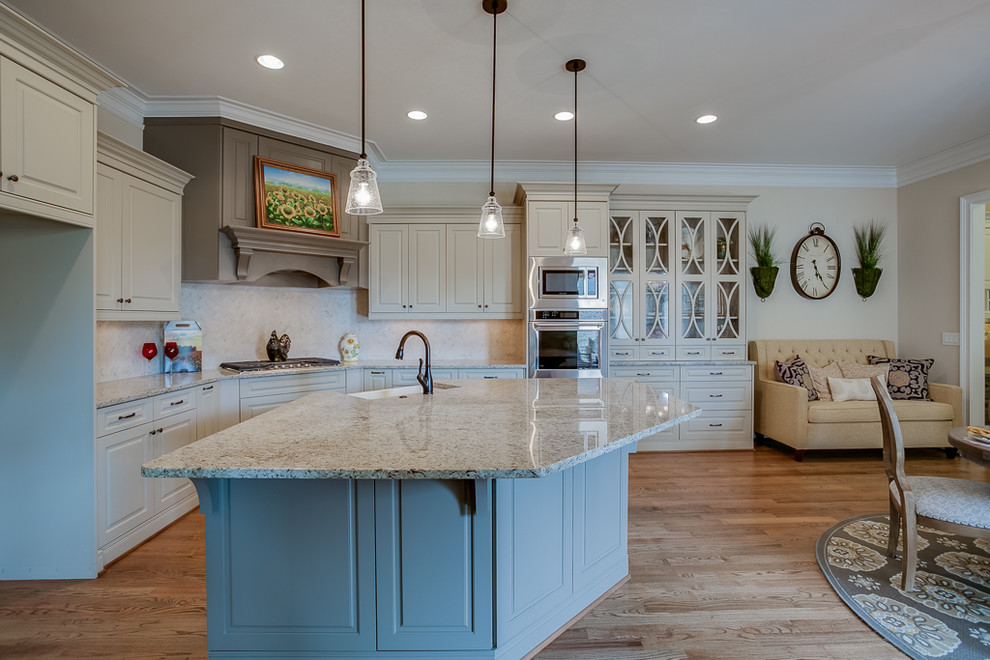 This is an example of a large traditional u-shaped kitchen in Other with medium hardwood floors, brown floor, a farmhouse sink, raised-panel cabinets, beige cabinets, quartz benchtops, black splashback, travertine splashback, stainless steel appliances, with island and multi-coloured benchtop.