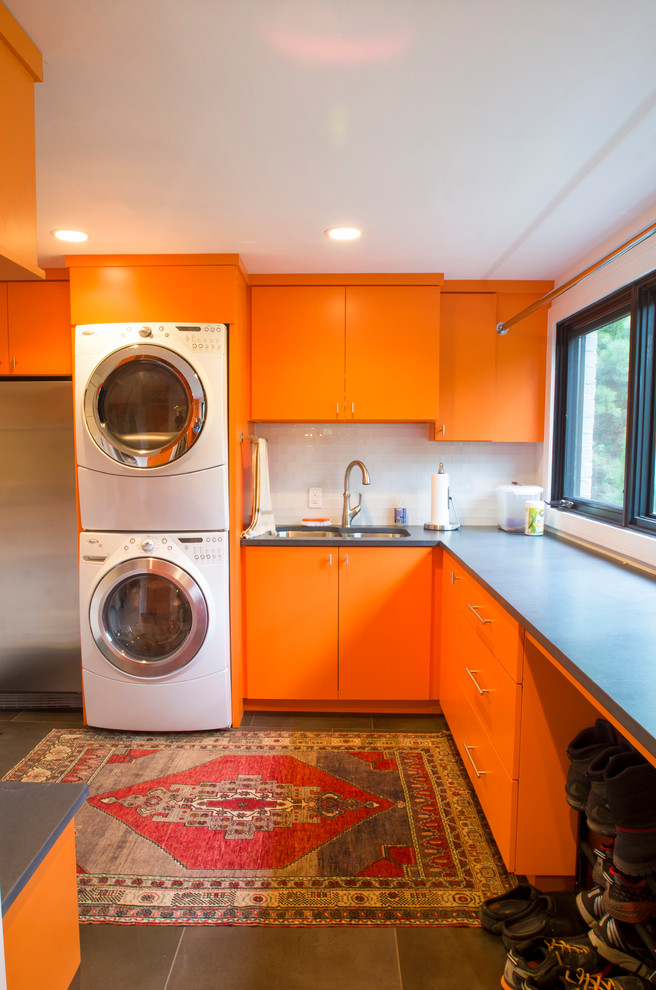 Inspiration for a large modern l-shaped laundry room in Milwaukee with an undermount sink, flat-panel cabinets, orange cabinets, quartzite benchtops, white walls, porcelain floors, a stacked washer and dryer, black floor and black benchtop.