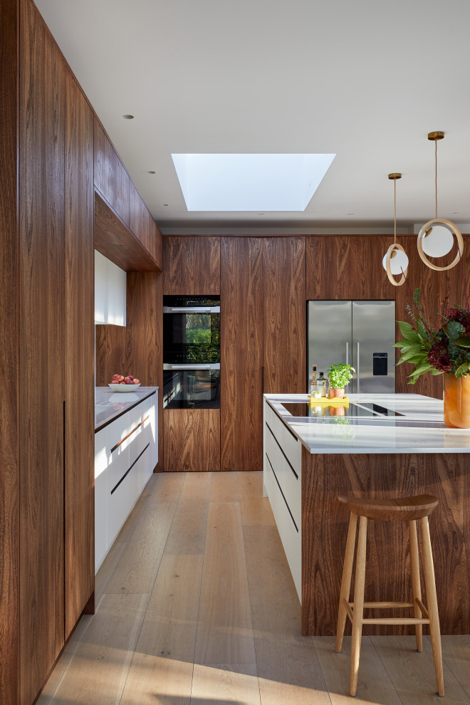 This is an example of a large transitional galley open plan kitchen in London with a double-bowl sink, flat-panel cabinets, medium wood cabinets, solid surface benchtops, white splashback, engineered quartz splashback, black appliances, medium hardwood floors, with island, brown floor and white benchtop.