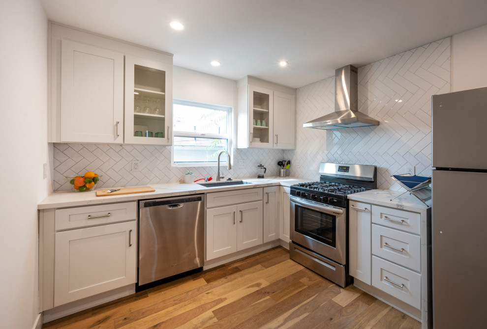 Mid-sized modern l-shaped eat-in kitchen in Los Angeles with a single-bowl sink, shaker cabinets, white cabinets, quartz benchtops, white splashback, cement tile splashback, stainless steel appliances, medium hardwood floors, no island, brown floor and white benchtop.