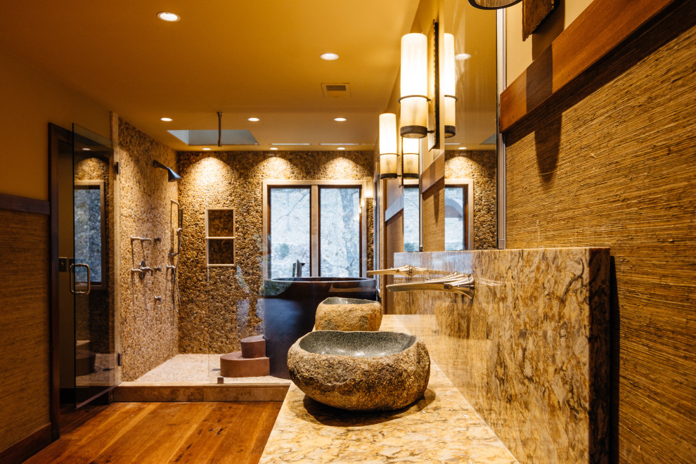 Photo of a modern master bathroom in Nashville with furniture-like cabinets, medium wood cabinets, a japanese tub, a shower/bathtub combo, a wall-mount toilet, gray tile, pebble tile, grey walls, medium hardwood floors, a vessel sink, marble benchtops, brown floor, a hinged shower door, yellow benchtops, a niche, a double vanity, a built-in vanity and decorative wall panelling.