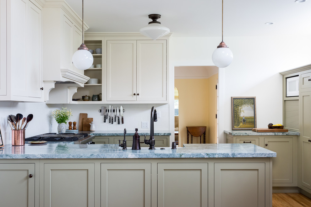 Small arts and crafts u-shaped kitchen in Los Angeles with a farmhouse sink, shaker cabinets, green cabinets, quartzite benchtops, white splashback, ceramic splashback, medium hardwood floors, grey benchtop, stainless steel appliances, a peninsula and brown floor.