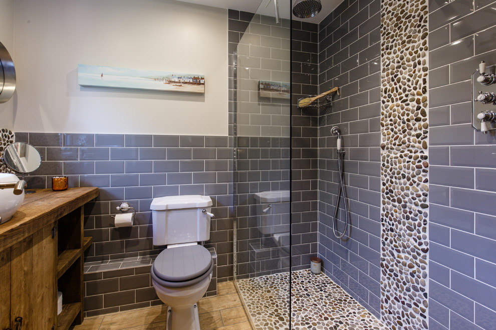 This is an example of a beach style master wet room bathroom in Cambridgeshire with dark wood cabinets, a one-piece toilet, gray tile, subway tile, grey walls, ceramic floors, a drop-in sink, wood benchtops, an open shower and flat-panel cabinets.