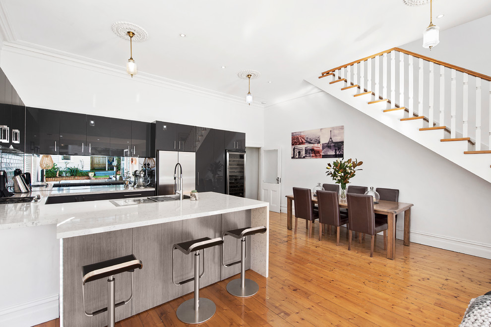 This is an example of a contemporary u-shaped open plan kitchen in Melbourne with a drop-in sink, flat-panel cabinets, black cabinets, glass sheet splashback, stainless steel appliances, light hardwood floors and a peninsula.