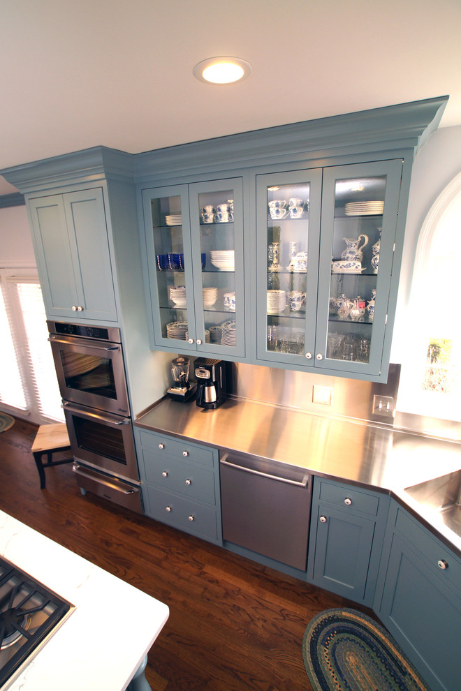 Design ideas for a large traditional u-shaped eat-in kitchen in St Louis with an integrated sink, blue cabinets, stainless steel benchtops, medium hardwood floors, with island, glass-front cabinets and metallic splashback.