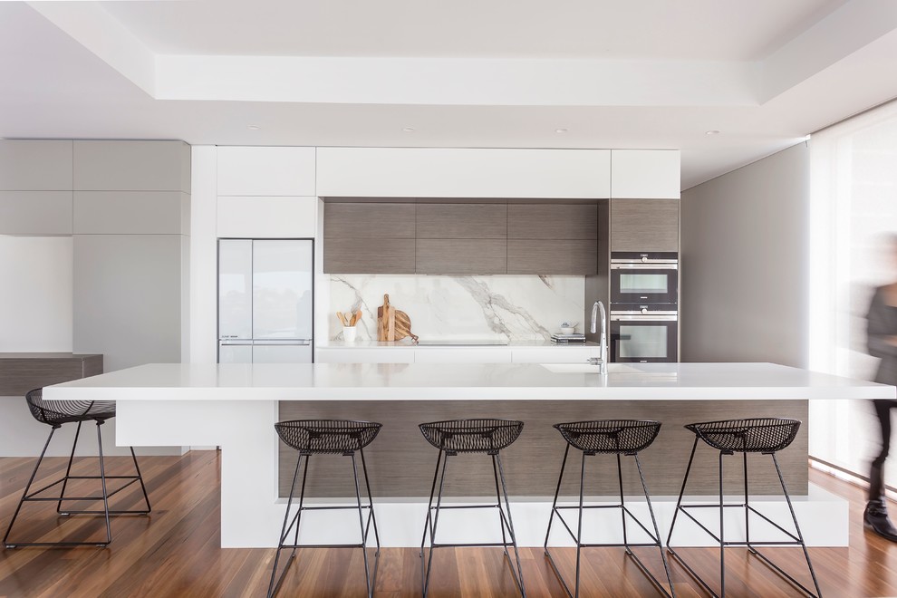 Design ideas for a large contemporary galley open plan kitchen in Sydney with an undermount sink, flat-panel cabinets, white cabinets, solid surface benchtops, white splashback, ceramic splashback, black appliances, light hardwood floors, with island, brown floor and white benchtop.