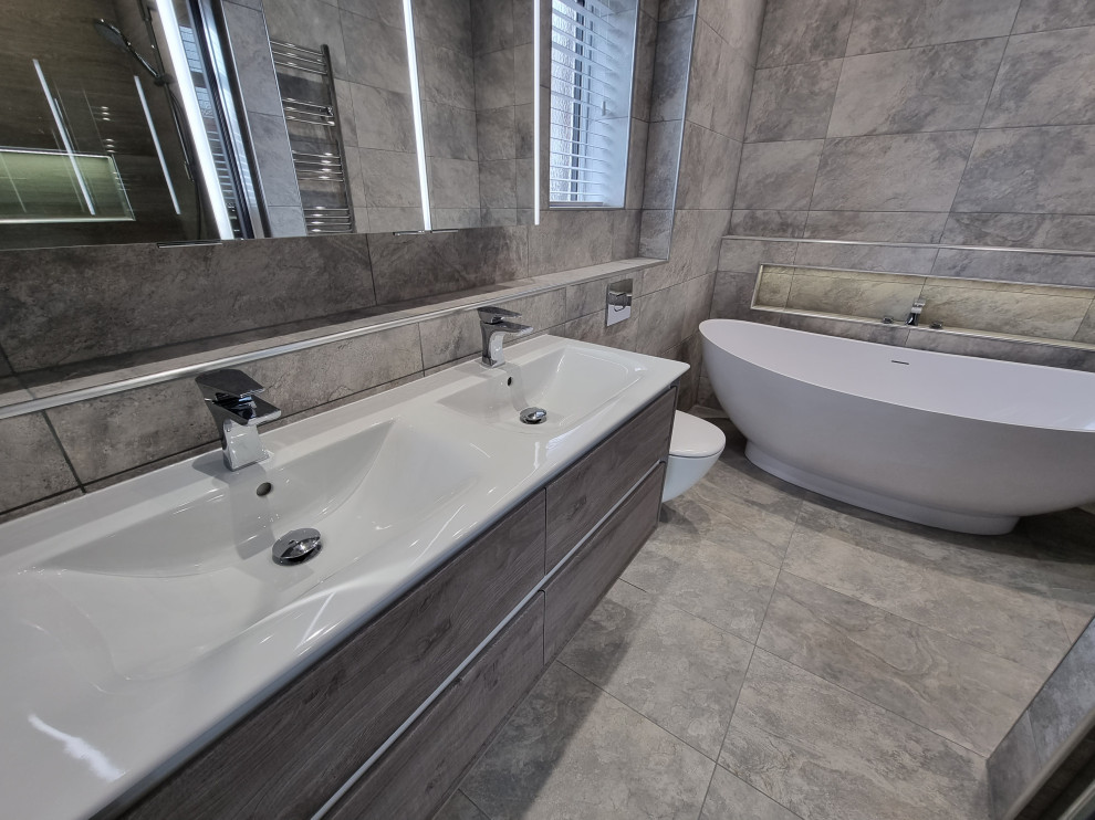 Photo of a large contemporary grey and brown ensuite bathroom in West Midlands with flat-panel cabinets, grey cabinets, a freestanding bath, a built-in shower, a wall mounted toilet, grey tiles, porcelain tiles, grey walls, porcelain flooring, a wall-mounted sink, grey floors, a hinged door, a wall niche, double sinks and a floating vanity unit.
