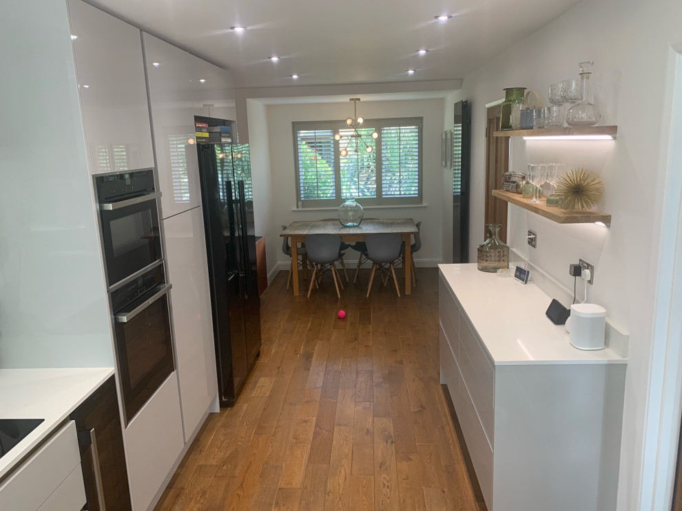 This is an example of a mid-sized contemporary l-shaped eat-in kitchen with flat-panel cabinets, grey cabinets, quartzite benchtops, white splashback, engineered quartz splashback, black appliances, no island, brown floor and white benchtop.