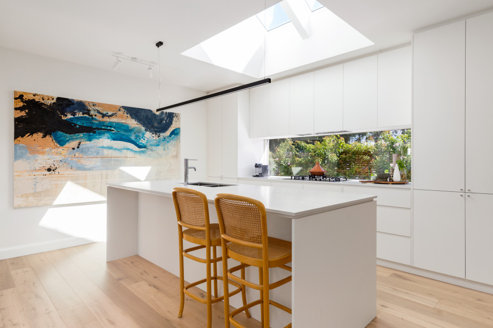 Inspiration for a mid-sized contemporary galley open plan kitchen in Melbourne with an undermount sink, flat-panel cabinets, white cabinets, quartz benchtops, window splashback, stainless steel appliances, light hardwood floors, with island, beige floor and white benchtop.