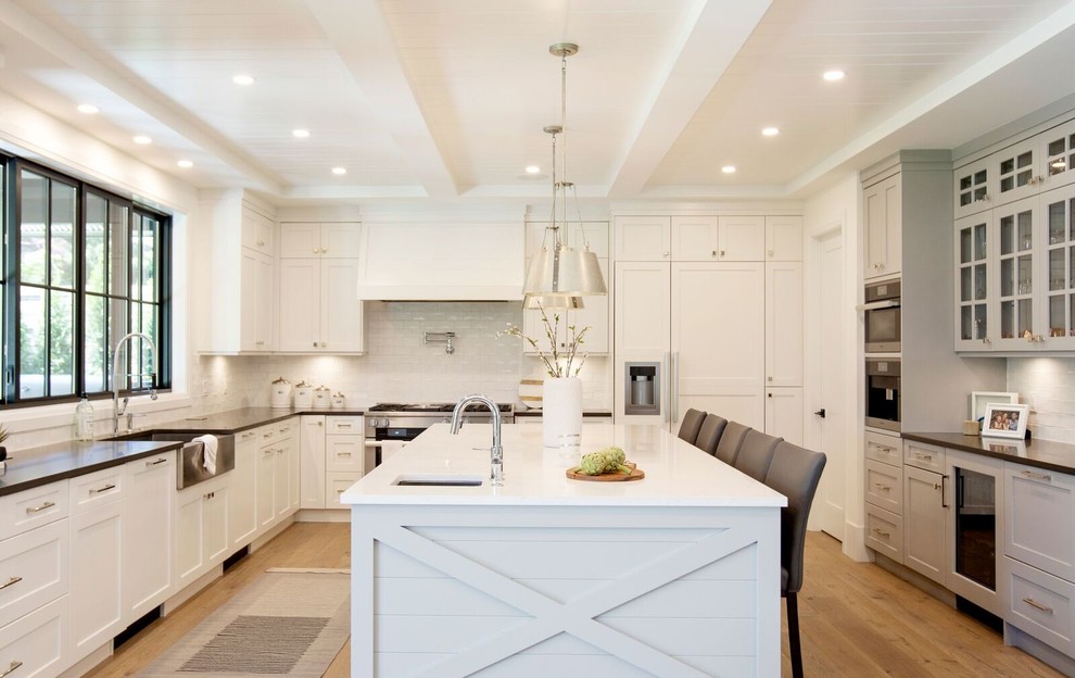 Photo of a large transitional u-shaped separate kitchen in Vancouver with a farmhouse sink, shaker cabinets, white cabinets, white splashback, subway tile splashback, stainless steel appliances, with island, black benchtop, light hardwood floors and brown floor.