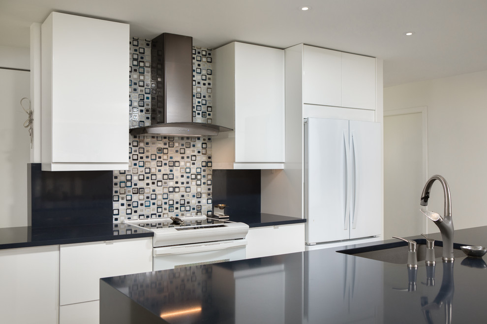Photo of a mid-sized contemporary l-shaped open plan kitchen in Miami with an undermount sink, flat-panel cabinets, white cabinets, solid surface benchtops, multi-coloured splashback, porcelain splashback, white appliances, light hardwood floors, with island, beige floor and black benchtop.