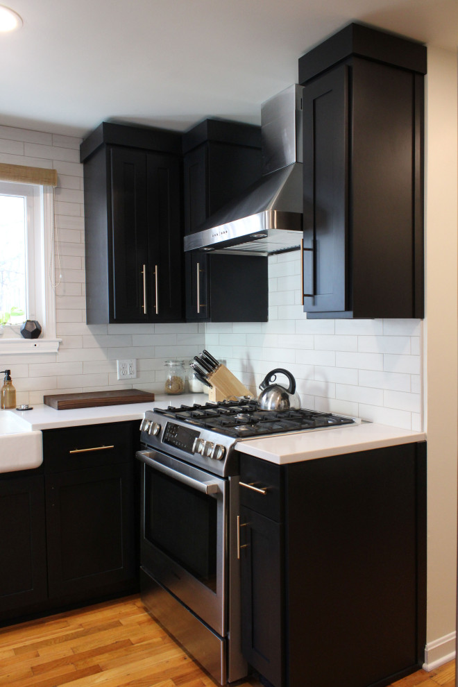This is an example of a small transitional l-shaped separate kitchen in Kansas City with a farmhouse sink, shaker cabinets, black cabinets, quartz benchtops, white splashback, subway tile splashback, stainless steel appliances, medium hardwood floors, no island and white benchtop.