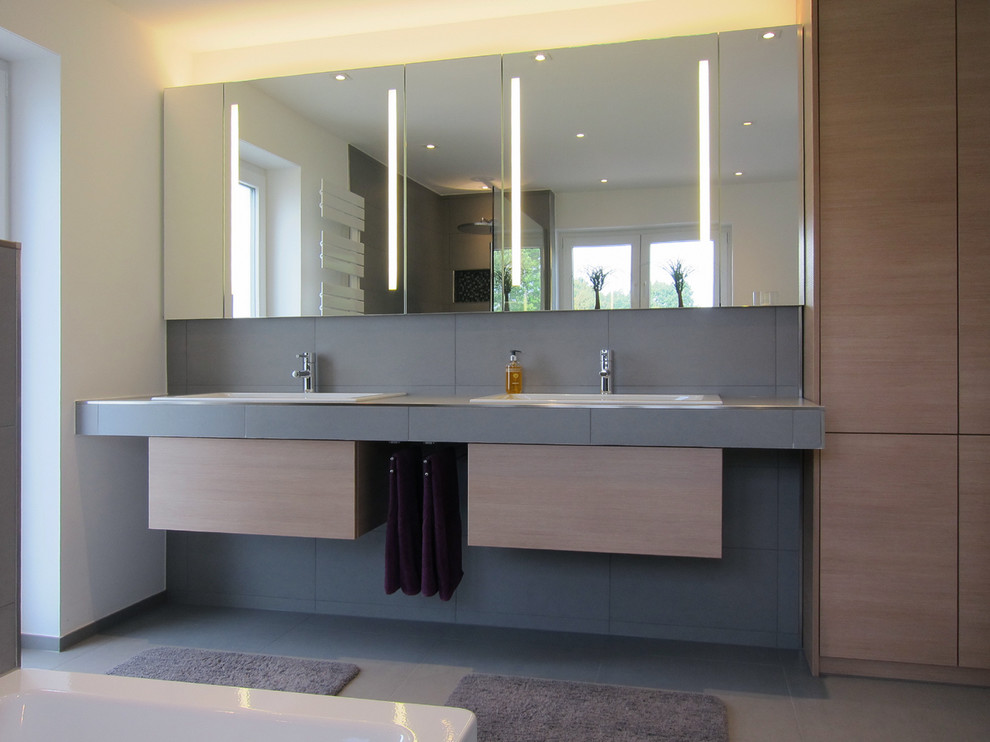 Badezimmer With Einbauwaschbecken, hellen Holzschränken, gefliestem Waschtisch, Einbaubadewanne, bodengleicher Dusche, grauen Fliesen, Keramikfliesen, weißer Wandfarbe, Keramikboden and flächenbündigen Schrankfronten in Cologne
