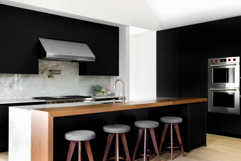 Large modern l-shaped open plan kitchen in Austin with a drop-in sink, flat-panel cabinets, black cabinets, marble benchtops, white splashback, marble splashback, stainless steel appliances, medium hardwood floors, with island, brown floor, white benchtop and recessed.