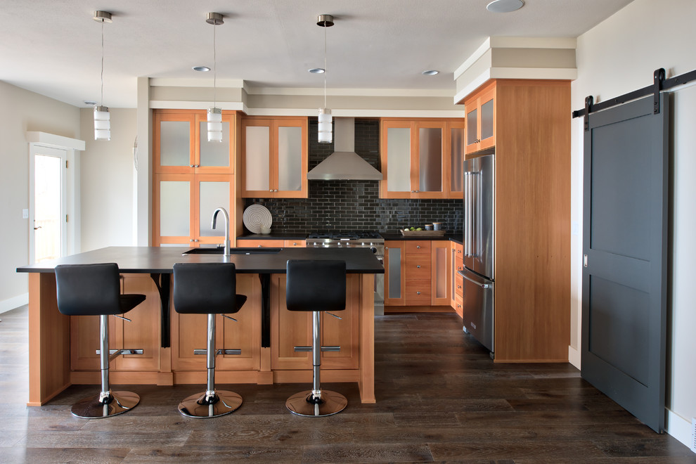 This is an example of a transitional l-shaped kitchen in Other with an undermount sink, recessed-panel cabinets, light wood cabinets, black splashback, subway tile splashback, stainless steel appliances, dark hardwood floors, with island, brown floor and black benchtop.